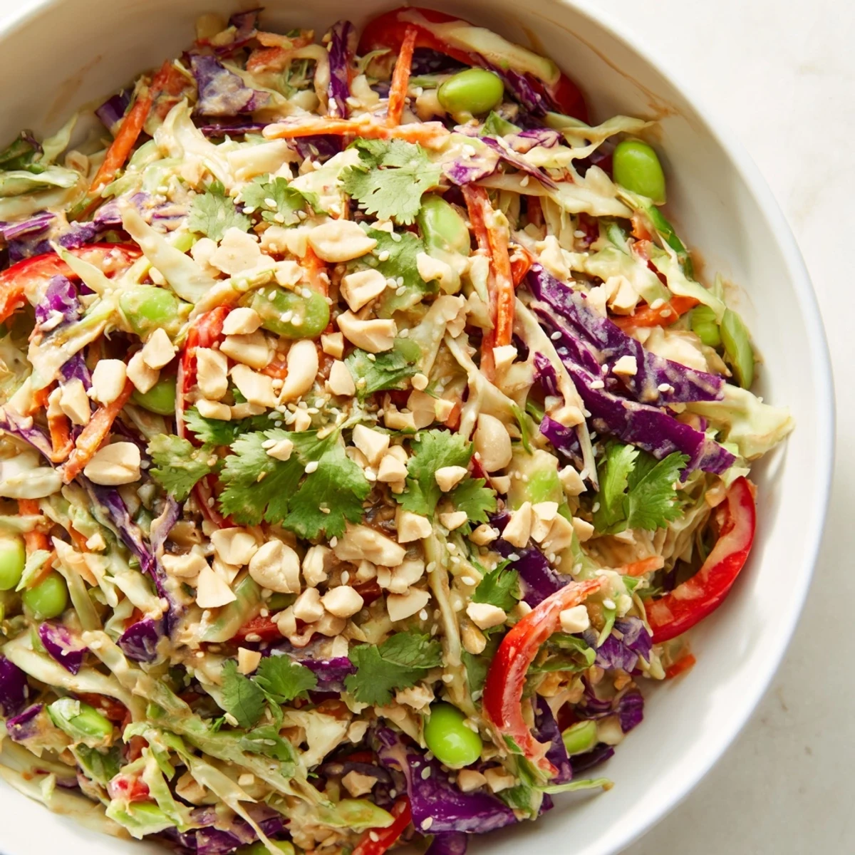 A colorful bowl of Crunchy Thai Peanut Salad with shredded carrots, purple cabbage, and edamame, drizzled with creamy peanut dressing and topped with chopped roasted peanuts for extra texture.