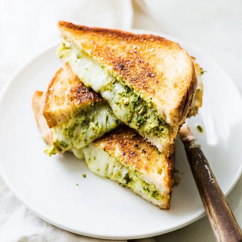Golden-brown sourdough pesto grilled cheese slices on a wooden board, revealing melted mozzarella, fontina, and cheddar inside.